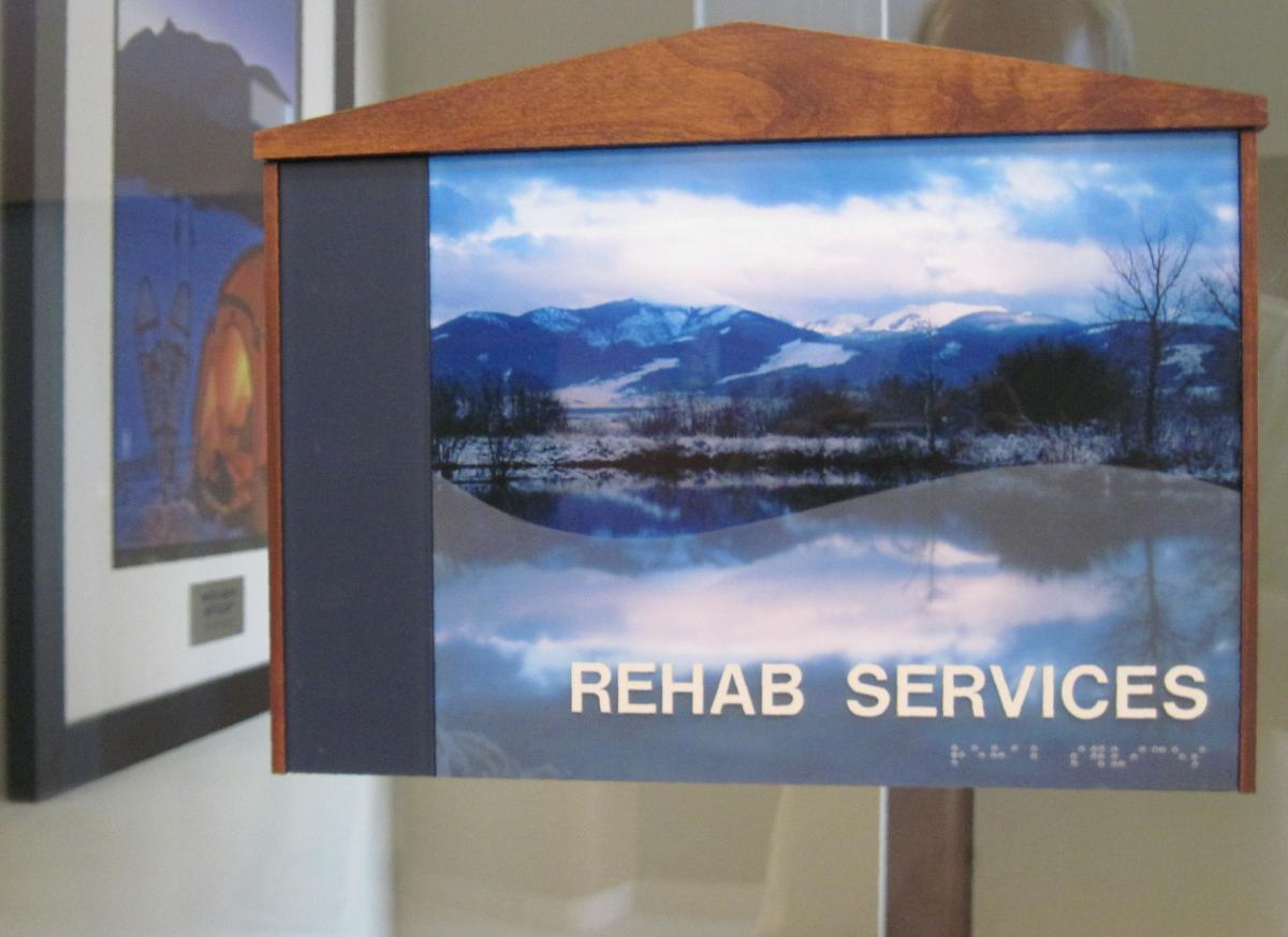 Sign for "Rehab Services" with a scenic photo of mountains and a lake. The sign has a wooden top and Braille beneath the text. The background features framed pictures on the wall.
