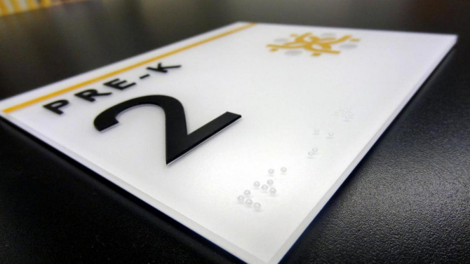A close-up view of a rectangular white sign on a dark surface. The sign features the text "PRE-K" in black, followed by the number "2" in large black print. There is also braille in the bottom left corner.