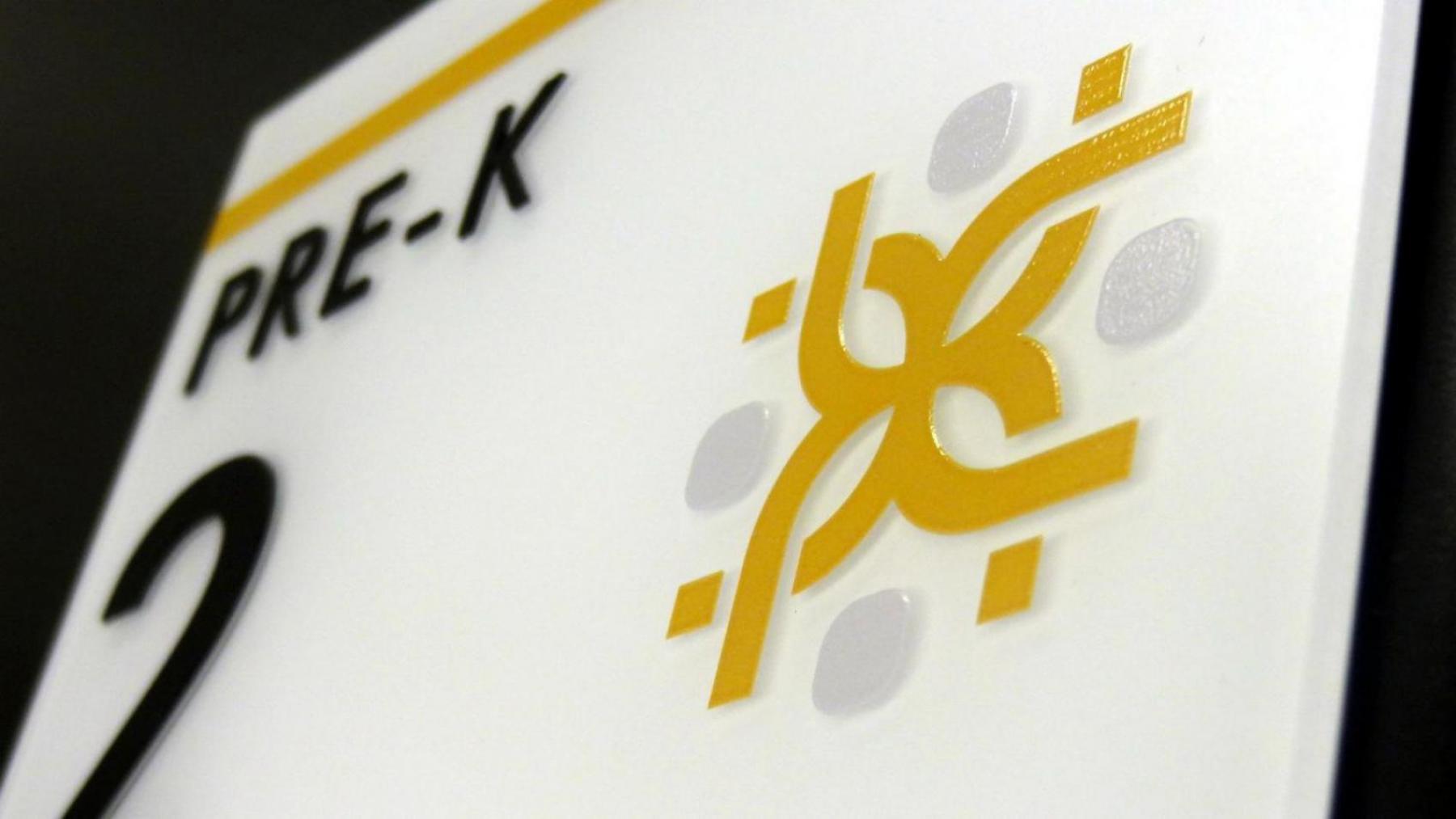 A close-up view of a rectangular white sign on a dark surface. The sign features the text "PRE-K" in black, followed by the number "2" in large black print. There is also braille in the bottom left corner.