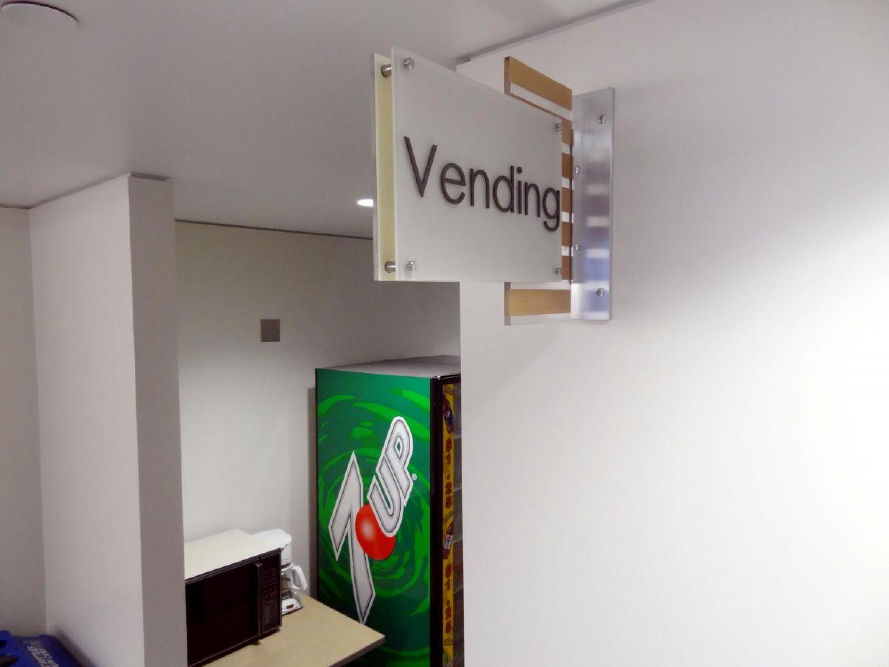 A vending area with a sign that reads "Vending" mounted on the wall. Below the sign, there is a green 7UP vending machine with a red button design. A microwave oven and a countertop are partially visible next to the vending machine.