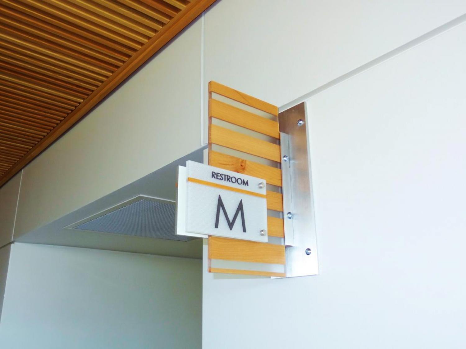 A restroom sign with a modern design is mounted on a wall. The sign has “RESTROOM” and the letter “M” indicating it is a men's restroom. The mount is made of wood and metal, and the background features a section of a ceiling with wooden slats.