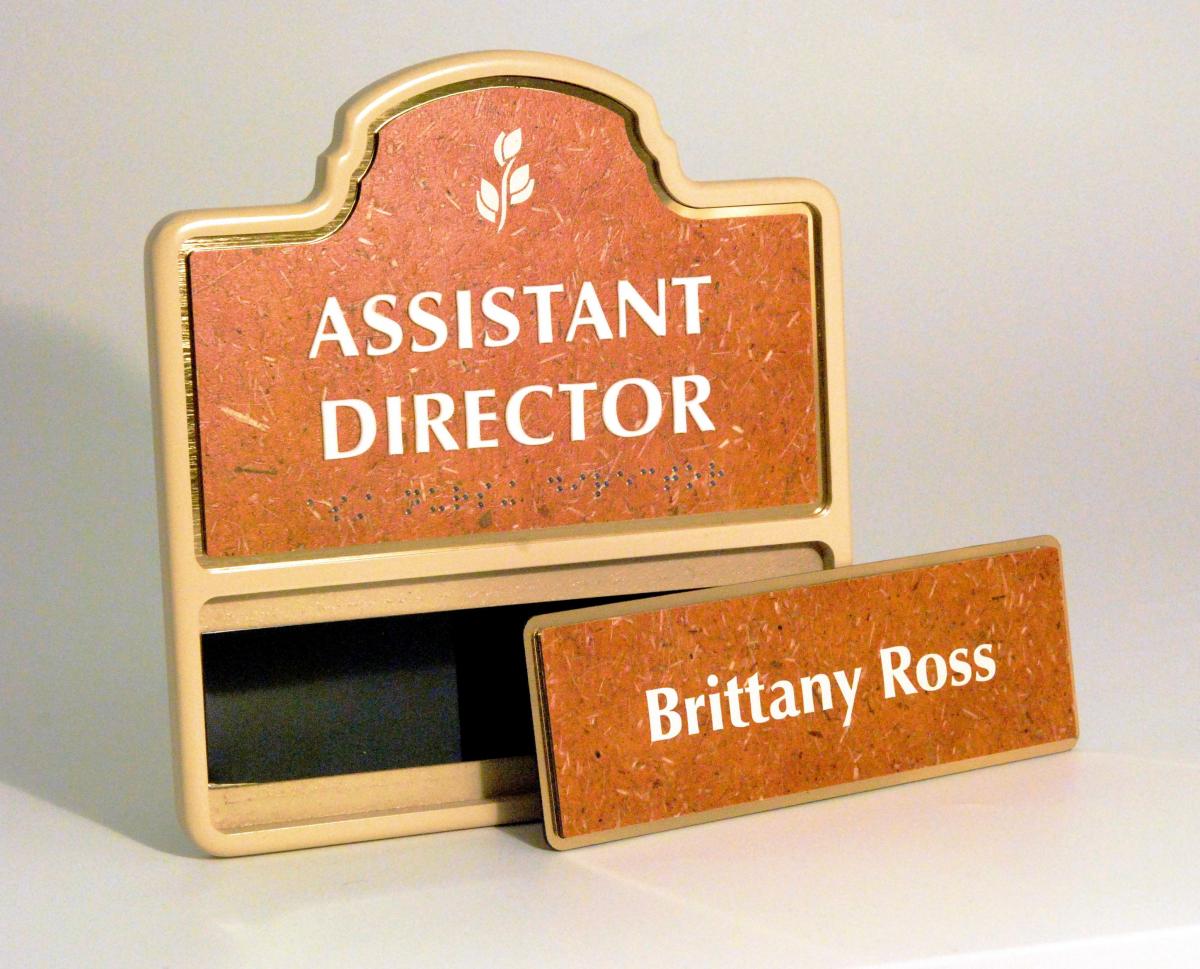 A wooden office nameplate holder with a carved design at the top, labeled "Assistant Director". Beneath it is a removable nameplate that reads "Brittany Ross". The holder and nameplate have a natural, corkboard-like finish.