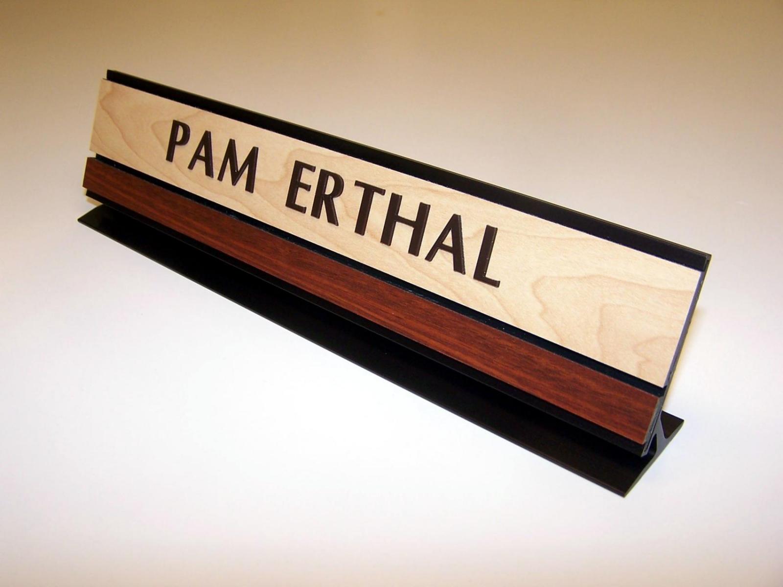 A wood and metal nameplate sits on a desk. The name "PAM ERTHAL" is engraved on the light wood section of the nameplate, with a dark wood strip beneath it. The base of the nameplate is black metal.