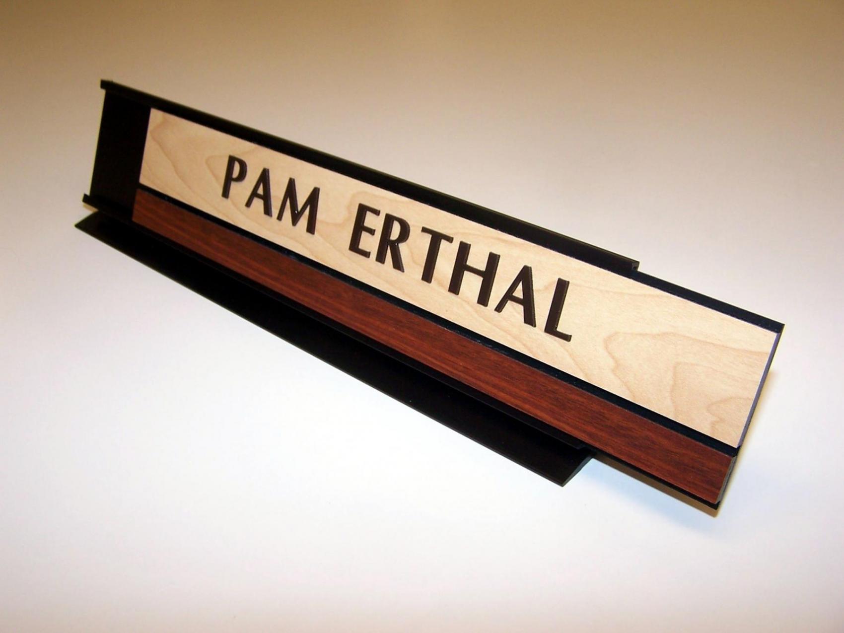 A wood and metal nameplate sits on a desk. The name "PAM ERTHAL" is engraved on the light wood section of the nameplate, with a dark wood strip beneath it. The base of the nameplate is black metal and the nameplate is half slid out.