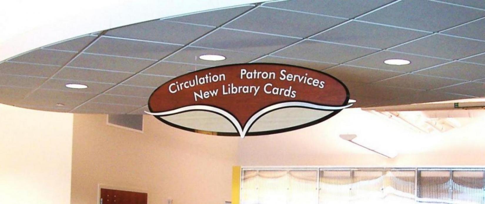 A ceiling-mounted sign in a library. The sign has a curved, book-like design and reads "Circulation, Patron Services, New Library Cards." The background shows part of the library's interior, including windows and lighting fixtures.