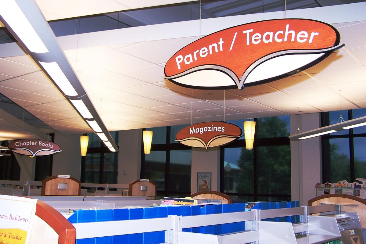 A library section with large overhead signs indicating different areas: "Parent/Teacher," "Chapter Books," and "Magazines." The area is well-lit with hanging pendant lights and features bookshelves and reading materials below the signs.