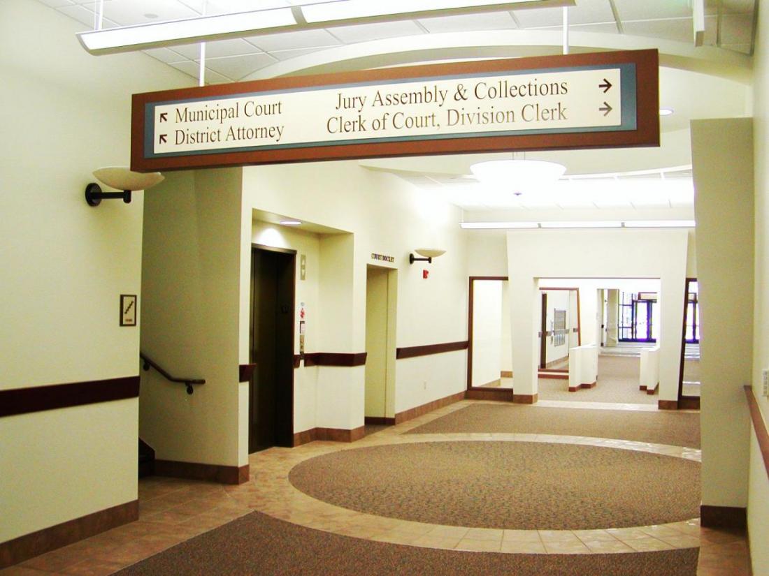 A bright hallway in a public building with beige walls and brown accents. Signs hang from the ceiling directing to Municipal Court, District Attorney, Jury Assembly & Collections, and Clerk of Court. Elevators and office doors are visible along the corridor.