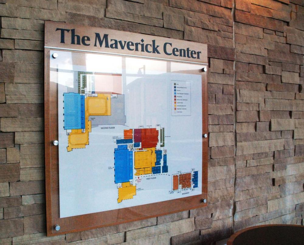 A framed floor plan of "The Maverick Center" is mounted on a stone wall. The map includes various colored sections representing different areas within the building. The background features a mix of brown and beige stone tiles.