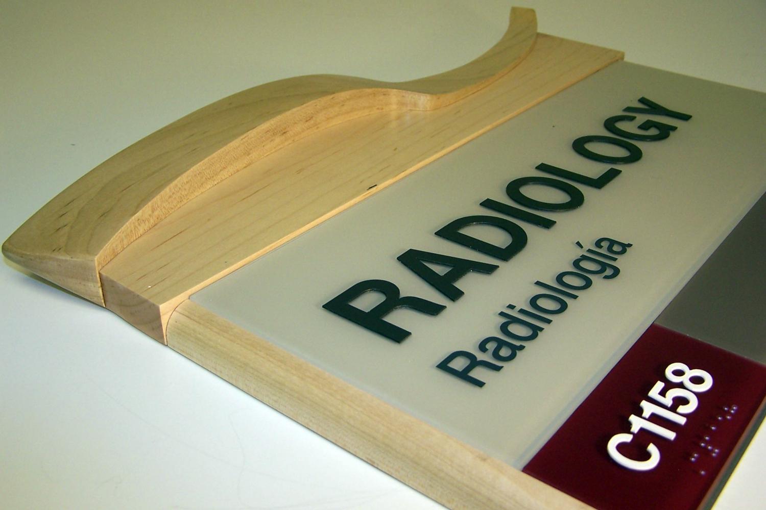 A close-up of a wall-mounted sign indicating a radiology department. The sign has text in both English ("RADIOLOGY") and Spanish ("Radiología"), along with the room number "C1158." The sign features a wooden frame with a curved decorative top.