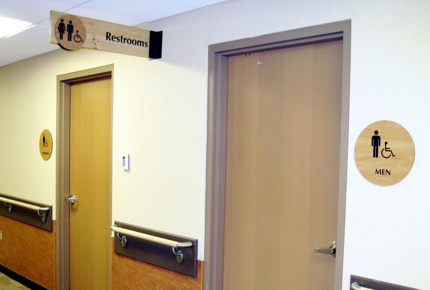 A hallway with two restroom doors. The left door has a sign for women, while the right door has a sign for men and accessibility. Above both doors is a hanging sign indicating the direction to the restrooms with symbols for men, women, and accessibility.