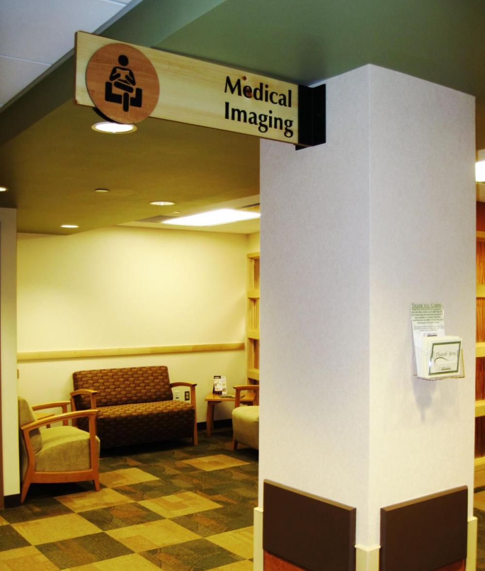 A hospital waiting area with beige walls and tiled flooring, featuring wooden chairs with plaid cushions. A sign overhead reads "Medical Imaging" and includes an icon of an individual sitting in front of a machine, directing patients to the imaging department.