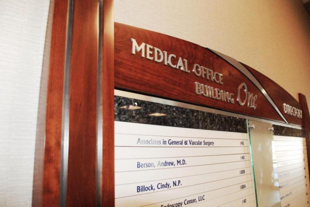 A wooden-framed directory on a wall titled "Medical Office Building One Directory." It lists offices and names of medical professionals, including their room numbers. The frame has a decorative arch at the top, and the names are displayed in columns.