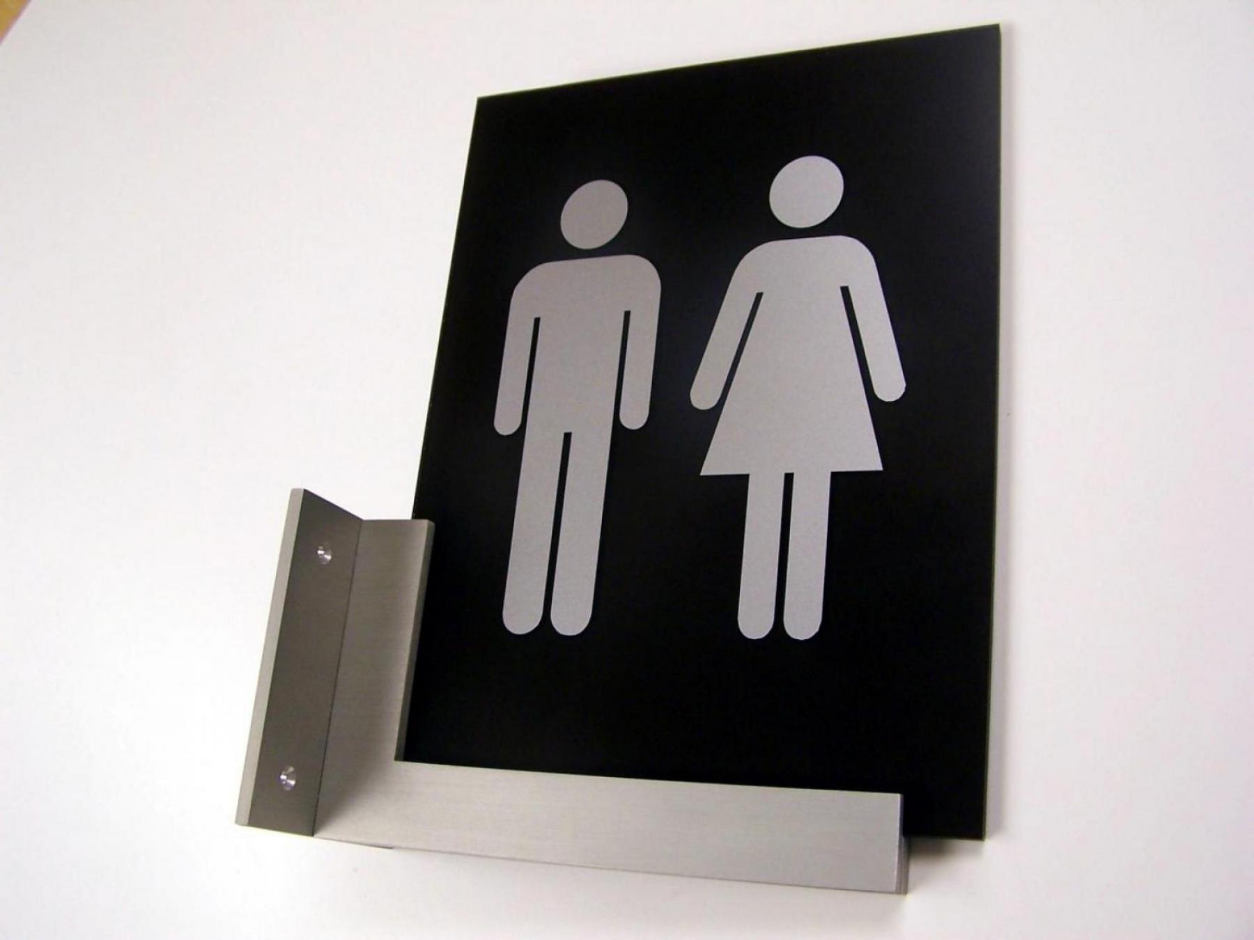 A rectangular bathroom sign mounted on a wall bracket with symbols indicating male and female restrooms. The sign features two simple white stick figures, one male and one female, on a dark background.