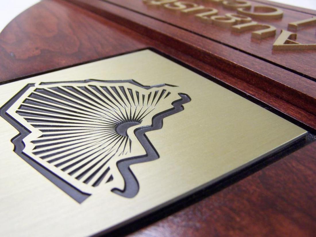 Close-up of a wooden plaque with a gold-colored metal plate on it. The plate features an engraved abstract design resembling the outline of a state with intricate lines radiating from its center. Part of an engraved inscription is visible along the top edge.