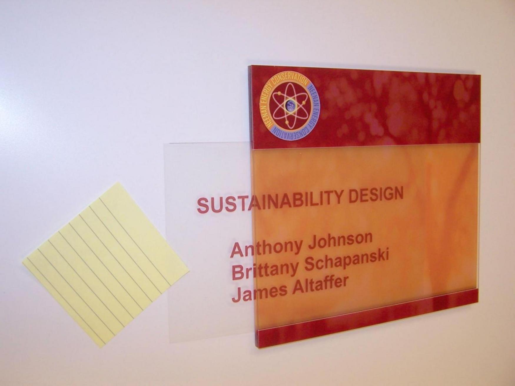 Plaque on a wall reading, "Sustainability Design" with names listed below: "Anthony Johnson, Brittany Schapanski, James Altaffer." The plaque has a red, marble-like background. The upper left corner features a logo with an atom design. There is a small yellow sticky note placed on the white wall to the left of the nameplate.