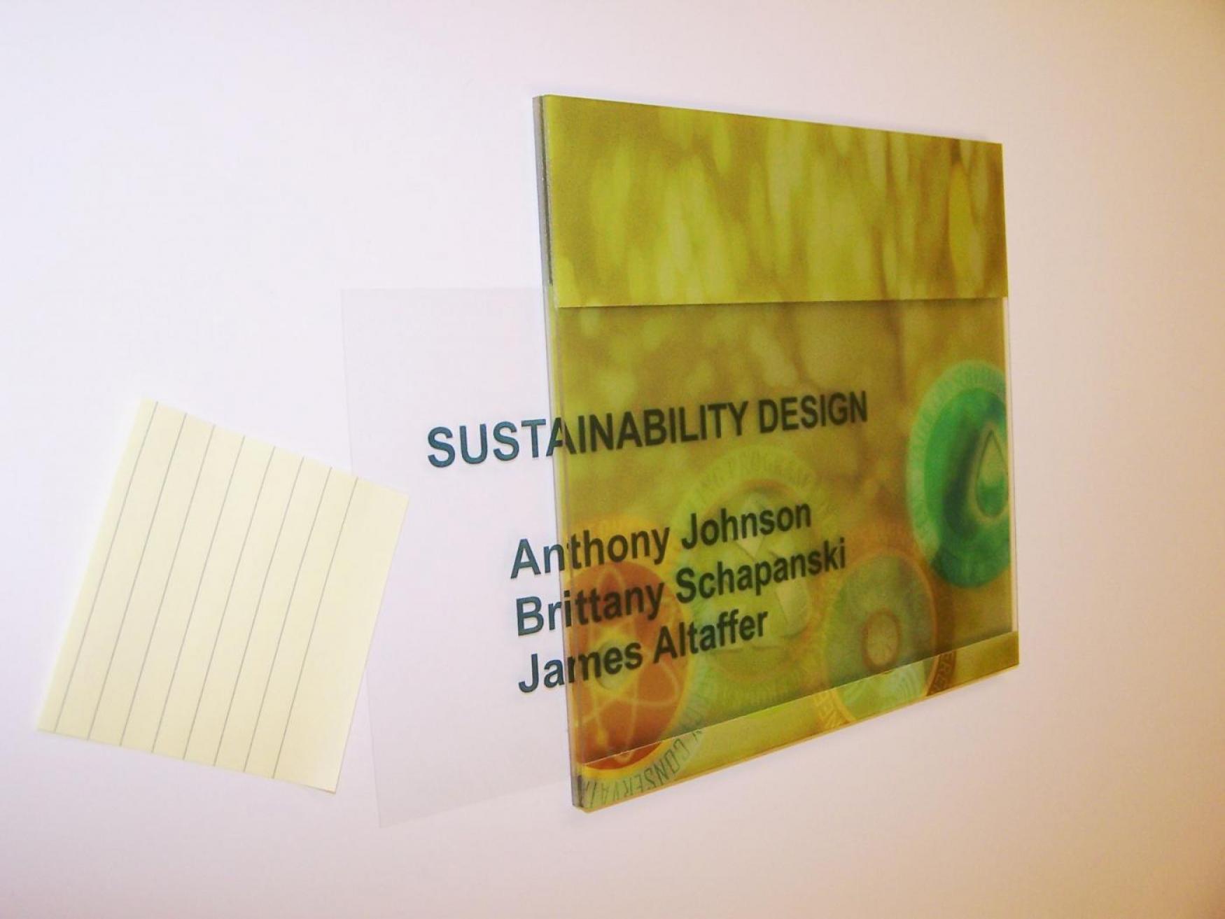 A plastic nameplate with the text "SUSTAINABILITY DESIGN" and the names "Anthony Johnson, Brittany Schapanski, James Altaffer" printed in bold. There is a small yellow sticky note placed on the white wall to the left of the nameplate.