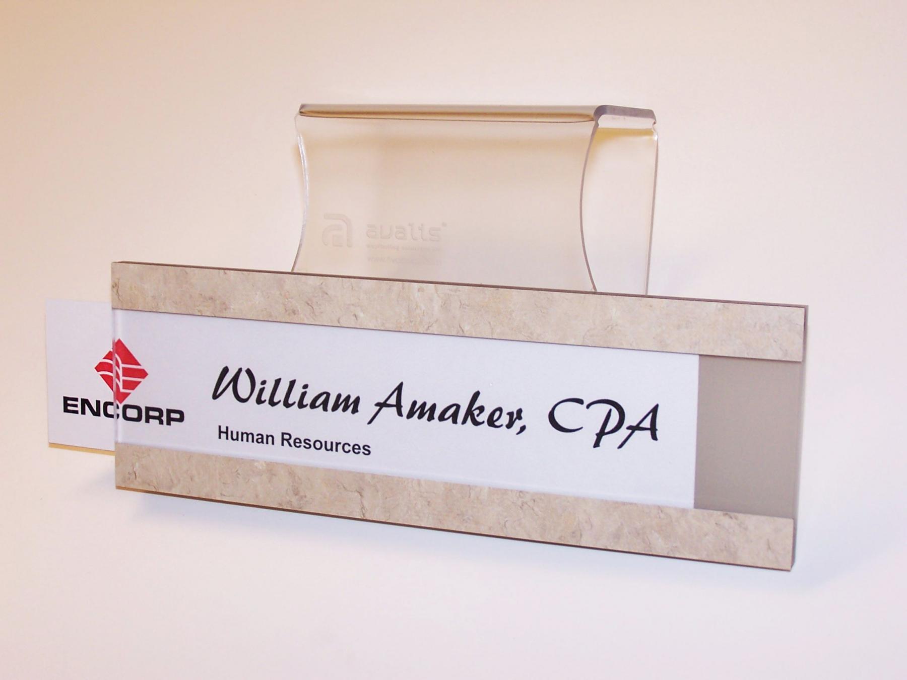 A desk nameplate holder displays a card reading "William Amaker, CPA, Human Resources." The holder is translucent with a silver frame. The card has a logo on the left side. A red and white business card with the Encorp logo is partially inserted behind the nameplate.
