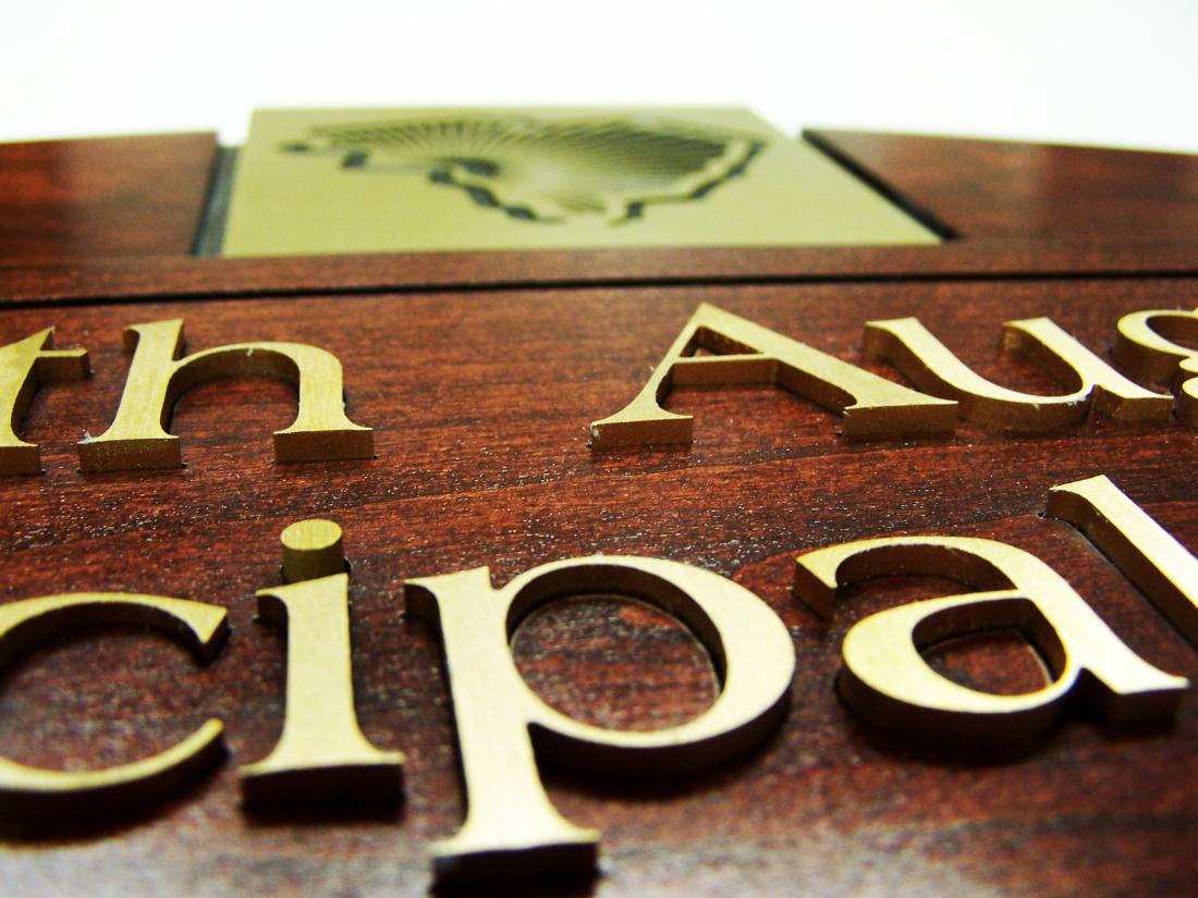 A close-up view of a wooden plaque with gold-colored text. The words are partially visible, showing "ncipal" and "th Aus" in clear focus. The background is blurred, highlighting the detailed texture and shimmer of the engraved letters.