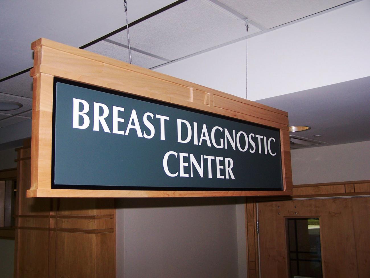 A rectangular wooden sign with a dark blue background and white text hangs from a ceiling, reading "BREAST DIAGNOSTIC CENTER." The sign is located indoors, with windows and fluorescent lights visible in the background.