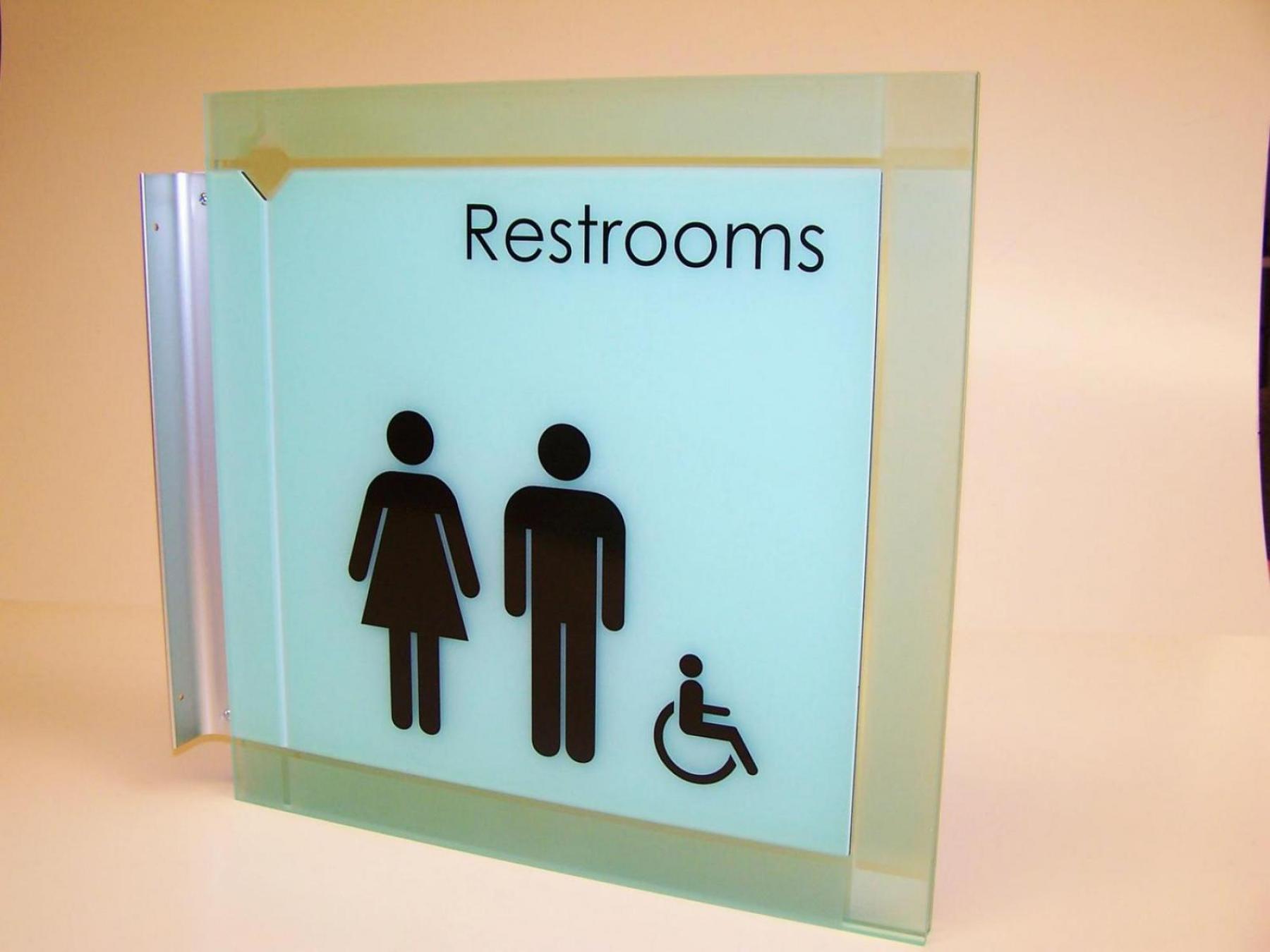 A close-up image of a frosted glass sign for restrooms, depicting symbols for male, female, and wheelchair accessibility. The sign is mounted on a wall, with another smaller sign below it that reads "Telluride Peak." The background is white.