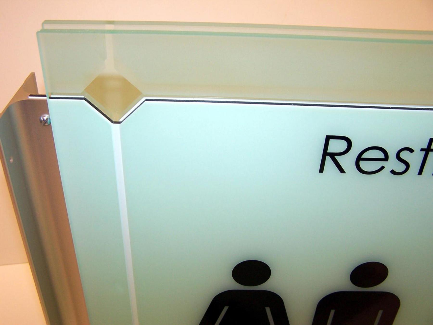 A close-up image of a frosted glass sign for restrooms, depicting symbols for male, female, and wheelchair accessibility. The sign is mounted on a wall. The background is white.