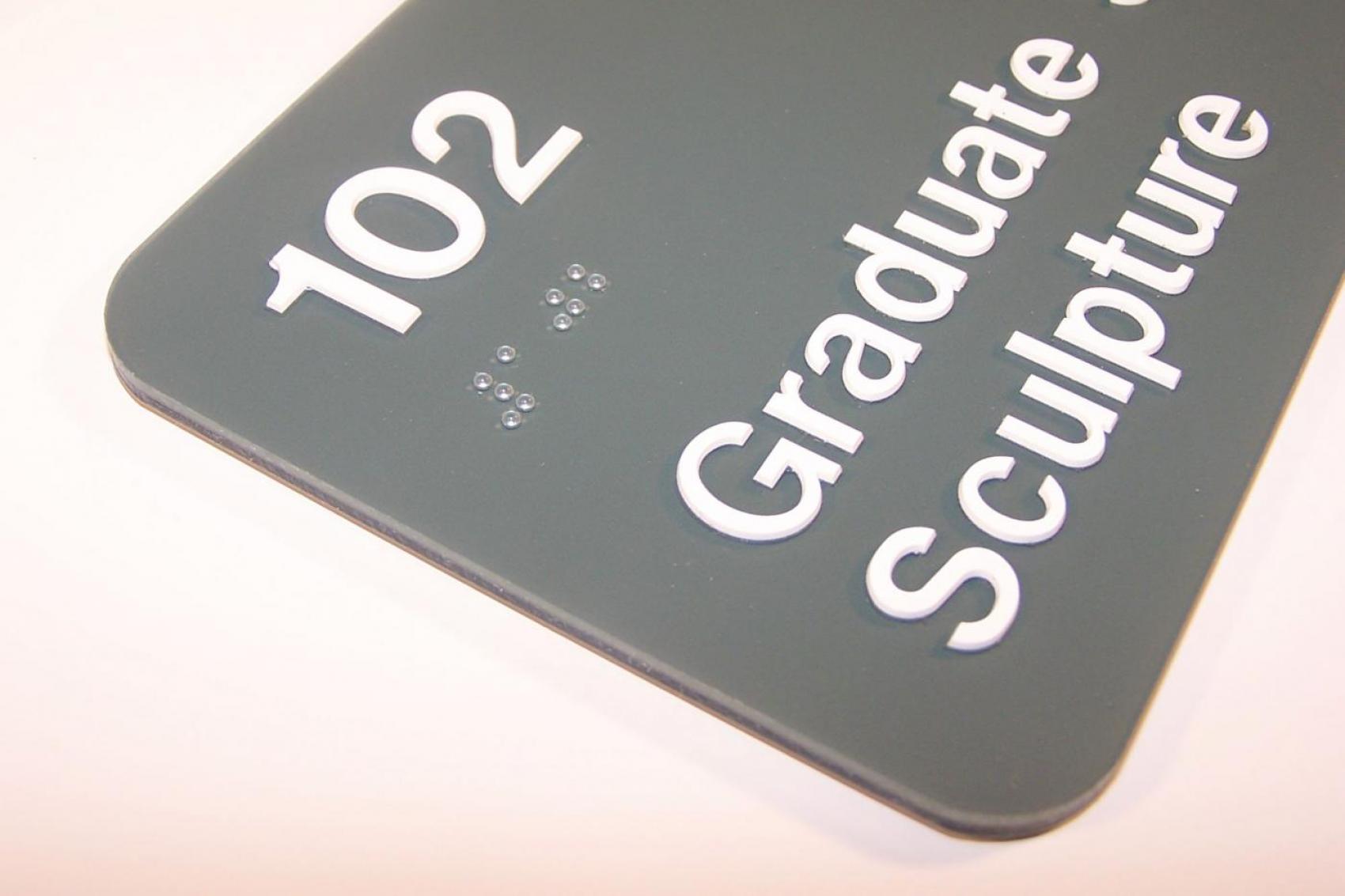 A dark grey tactile sign with the text "102 Graduate Sculpture" embossed in white. Braille dots representing the number "102" are also present below the number. The sign has rounded corners and is photographed against a light background.