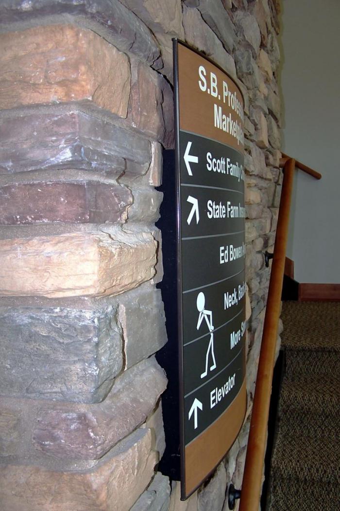A directional sign on a stone wall pointing to various offices and services including Scott Family, State Farm, Ed Bowen, Neck, More, and Elevator. The sign uses both text and simple stick-figure icons to indicate directions. A wooden railing is visible nearby.