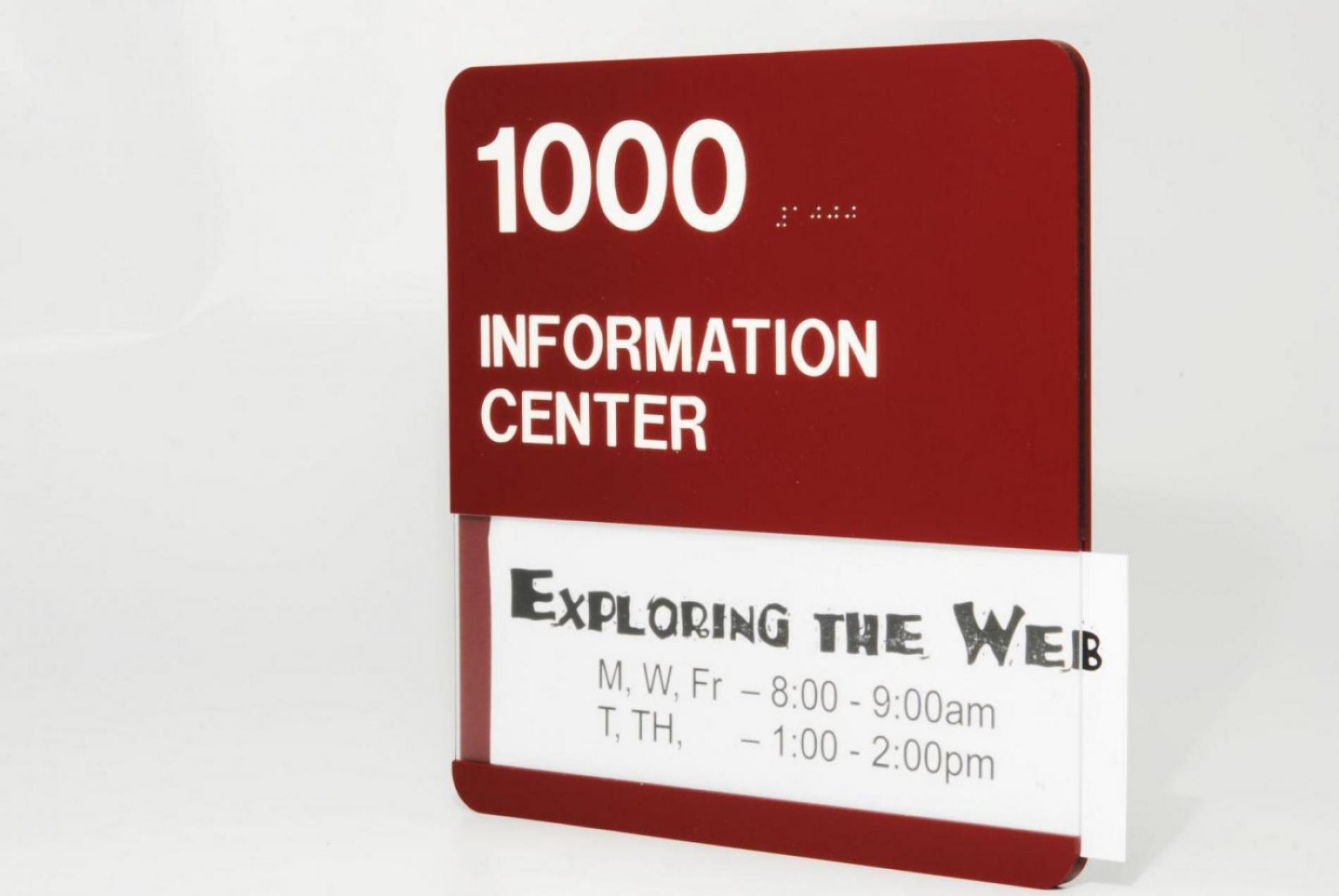 A red and white sign with "1000 INFORMATION CENTER" and Braille symbols at the top. Below is a holder with a paper labeled "Exploring the Web," showing class times: M, W, Fr from 8:00-9:00am and T, TH from 1:00-2:00pm.
