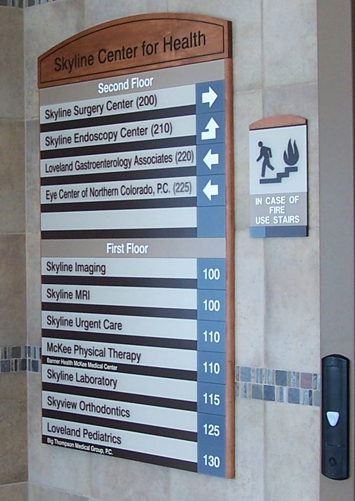 A directory sign in the Skyline Center for Health building. It lists departments on two floors: Surgery Center, Endoscopy Center, Gastroenterology Associates, and Eye Center on the second floor; Imaging, MRI, Urgent Care, Physical Therapy, Laboratory, and Pediatrics on the first floor. There's an adjacent sign showing a person using stairs with text instructing to use stairs in case of fire.