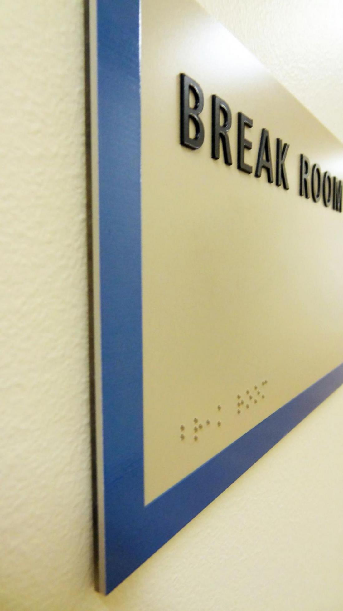 A close-up side view of a rectangular wall sign with blue borders. The sign reads "BREAK ROOM" in raised black letters and includes Braille script at the bottom. The sign is mounted on a white textured wall.