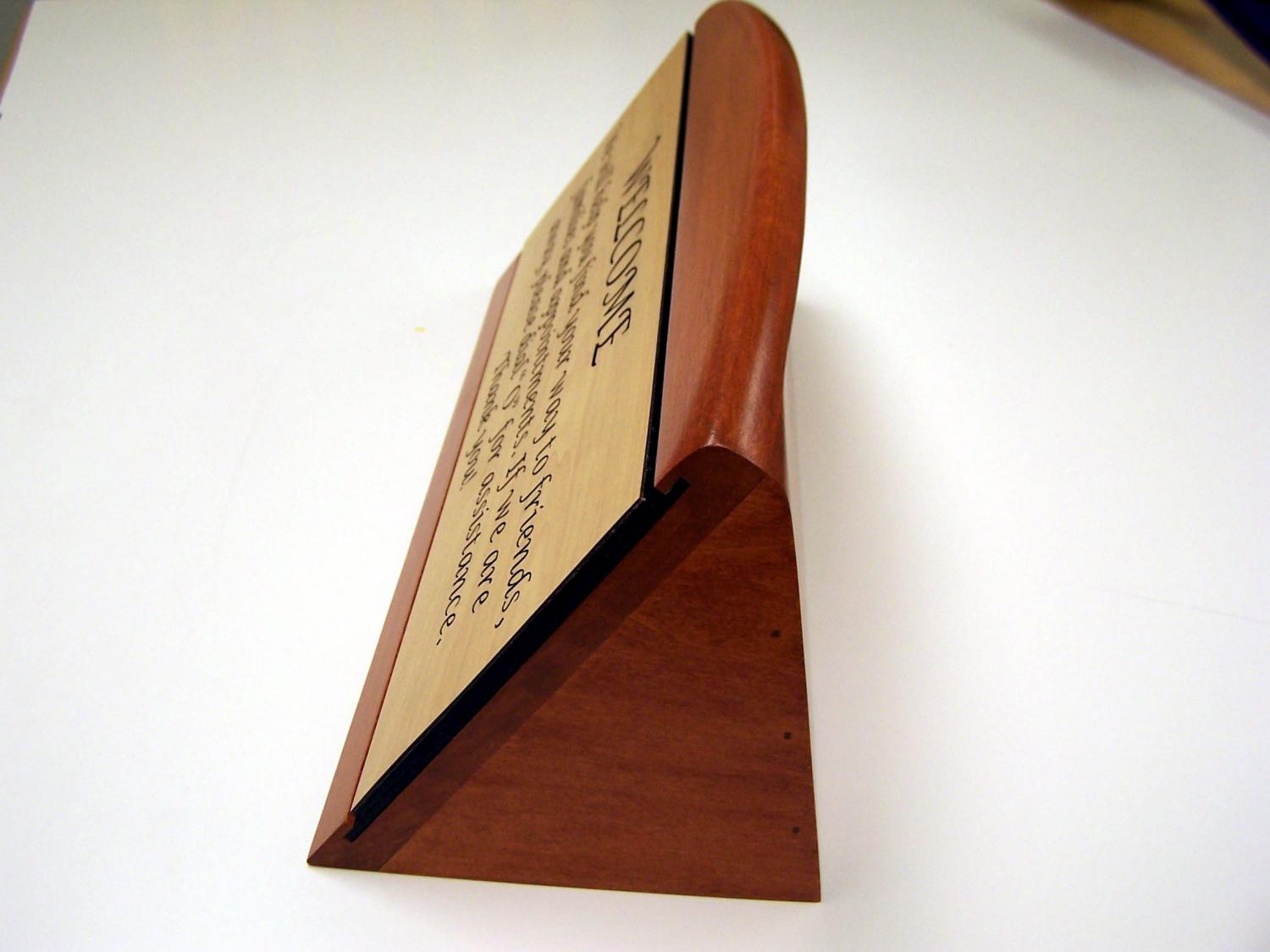 A wooden desk sign with a light-colored front panel engraved with a welcome message in elegant script. The message reads, "WELCOME. We will help you find your way to friends, families and appointments. If we are away, please dial '0' for assistance. Thank you!.  This is a side view closeup.