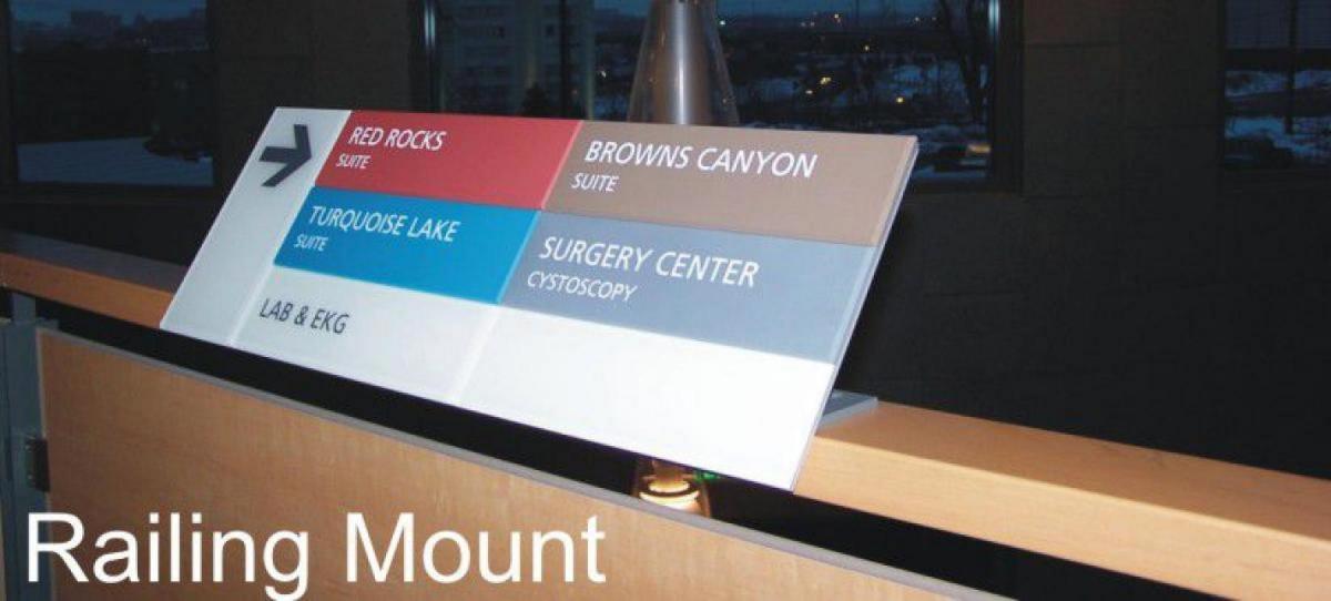 A directional sign mounted on a railing. The sign points to various locations within a building: Red Rocks Suite, Browns Canyon Suite, Turquoise Lake Suite, Surgery Center (Cystoscopy), and Lab & EKG. The background shows a dimly lit interior with windows.
