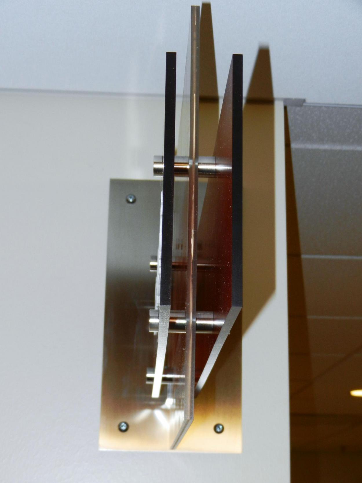 A close-up view of a modern, wall-mounted sign with two parallel glass panels held by metal bolts. The fixture is sleek and minimalist, installed on a beige wall, and lit by overhead lighting, casting subtle shadows on the wall.