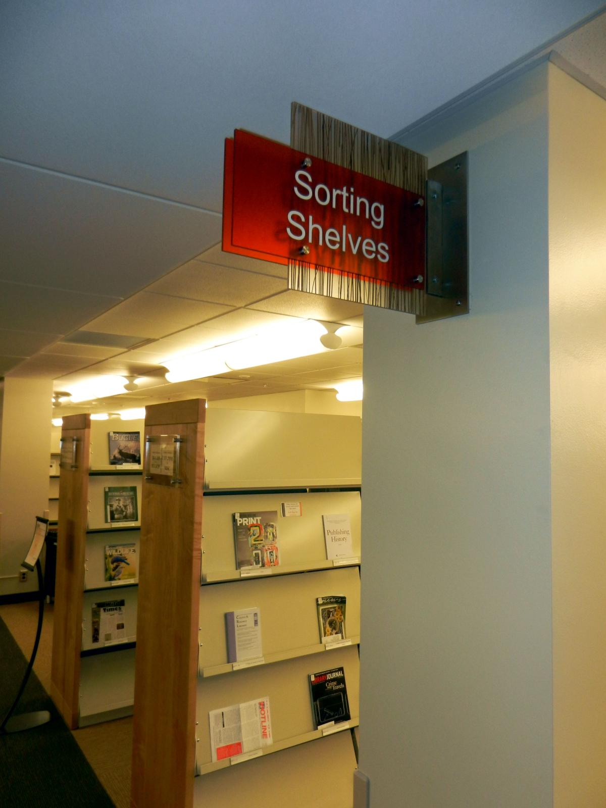 A library aisle with wooden shelves displaying various magazines and books. A red sign reading "Sorting Shelves" hangs from the ceiling at the entrance to the aisle. The area is well-lit with overhead lights and has a quiet, organized atmosphere.