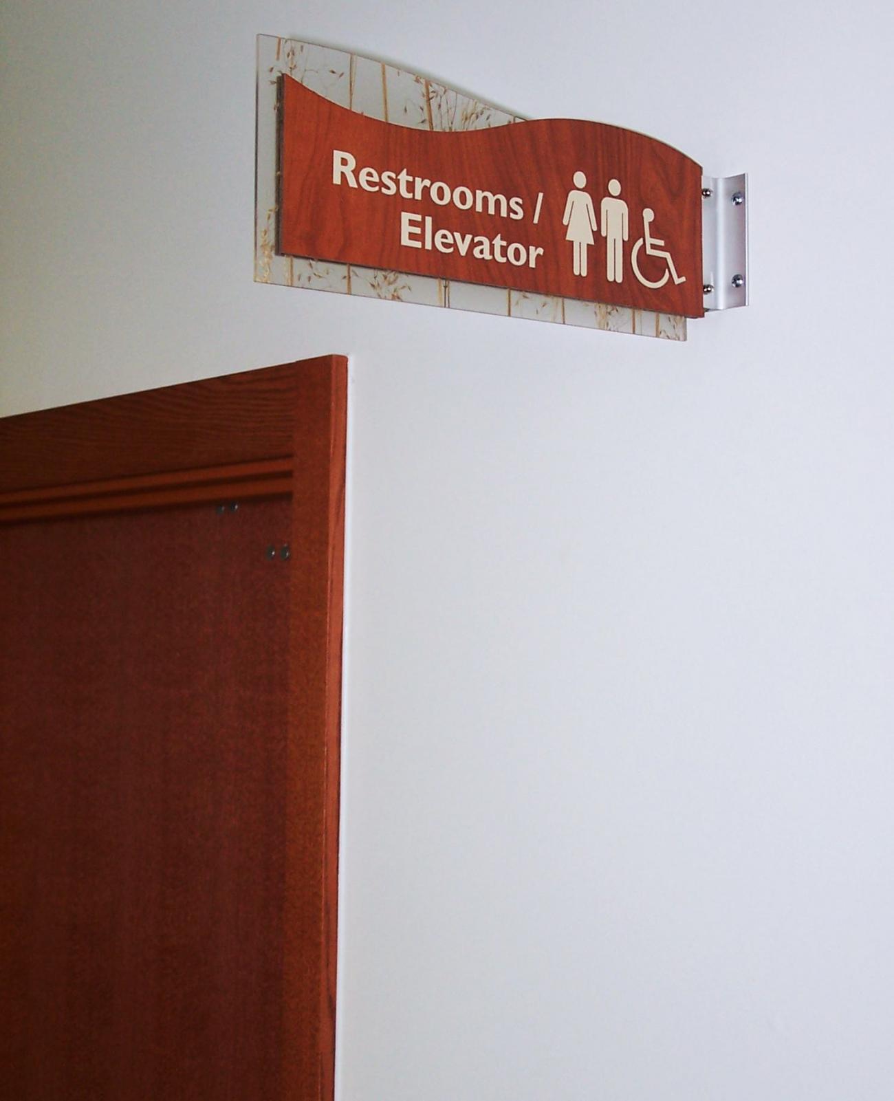 A wooden sign with white icons and text indicates the locations of restrooms and an elevator. The sign features symbols for men, women, and individuals with disabilities. The sign is mounted on a wall in a hallway.