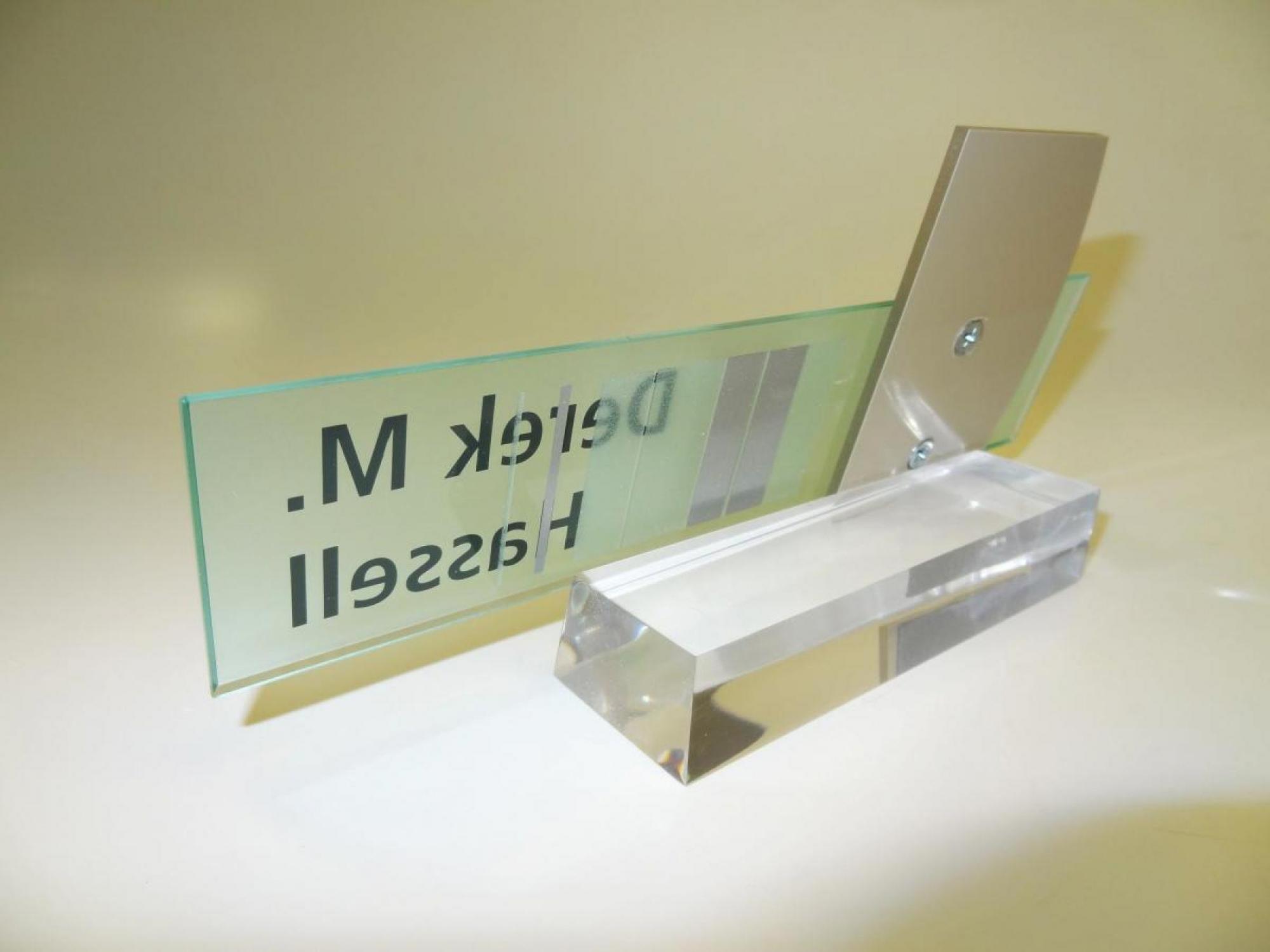 A glass nameplate with the text "Derek M. Hassell" engraved on it. The nameplate is mounted on a transparent, angled stand with a brushed metal bracket. The background is a plain light yellow surface.  This image shows the back of the nameplate.