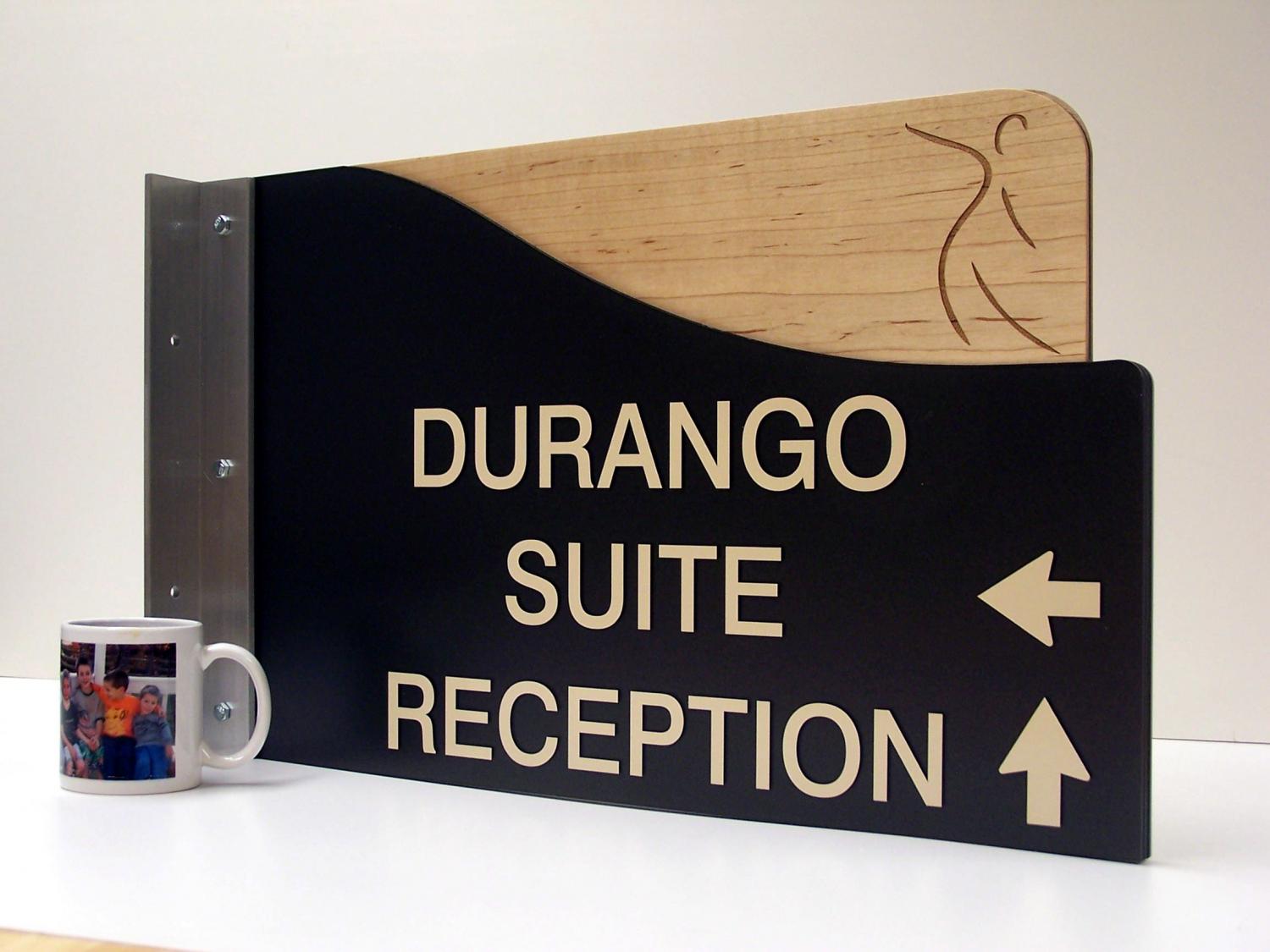 A black and wooden directional sign with a metal bracket. The sign reads "DURANGO SUITE" with an arrow pointing left and "RECEPTION" with an arrow pointing up. The top right corner features a subtle line drawing of a figure.  A coffee cup sits by the left side of the sign.