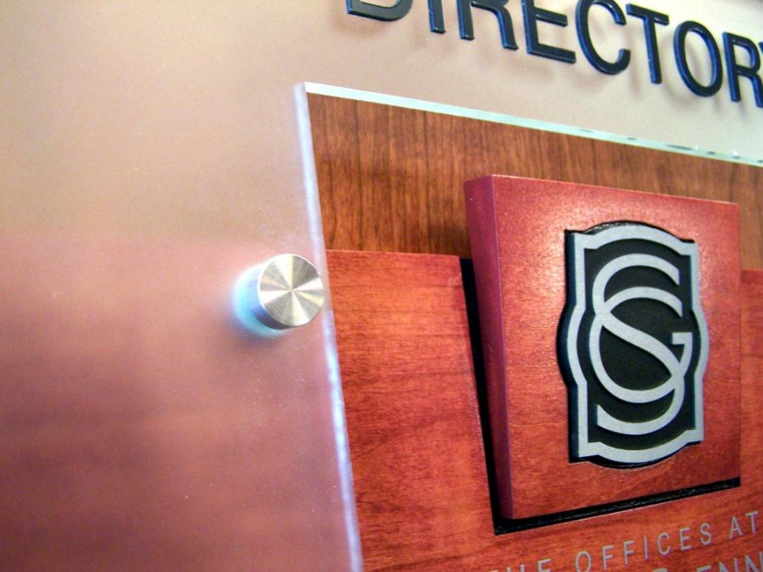 Close-up of a wall-mounted directory sign with a metallic edge and visible bolt. The sign features a black and white logo with intertwined "S" and "G" letters on a brown wooden background. Partial text "DIRECTORY" is visible at the top.