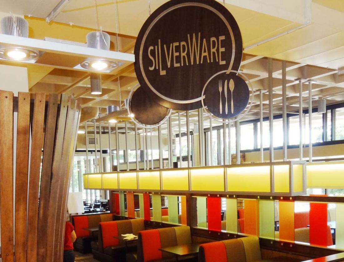 A dining area sign reading "SILVERWARE" hangs from the ceiling, featuring a fork, knife, and spoon icon. In the background are colorful tiles and a ceiling fixture designed with vertical wooden slats.