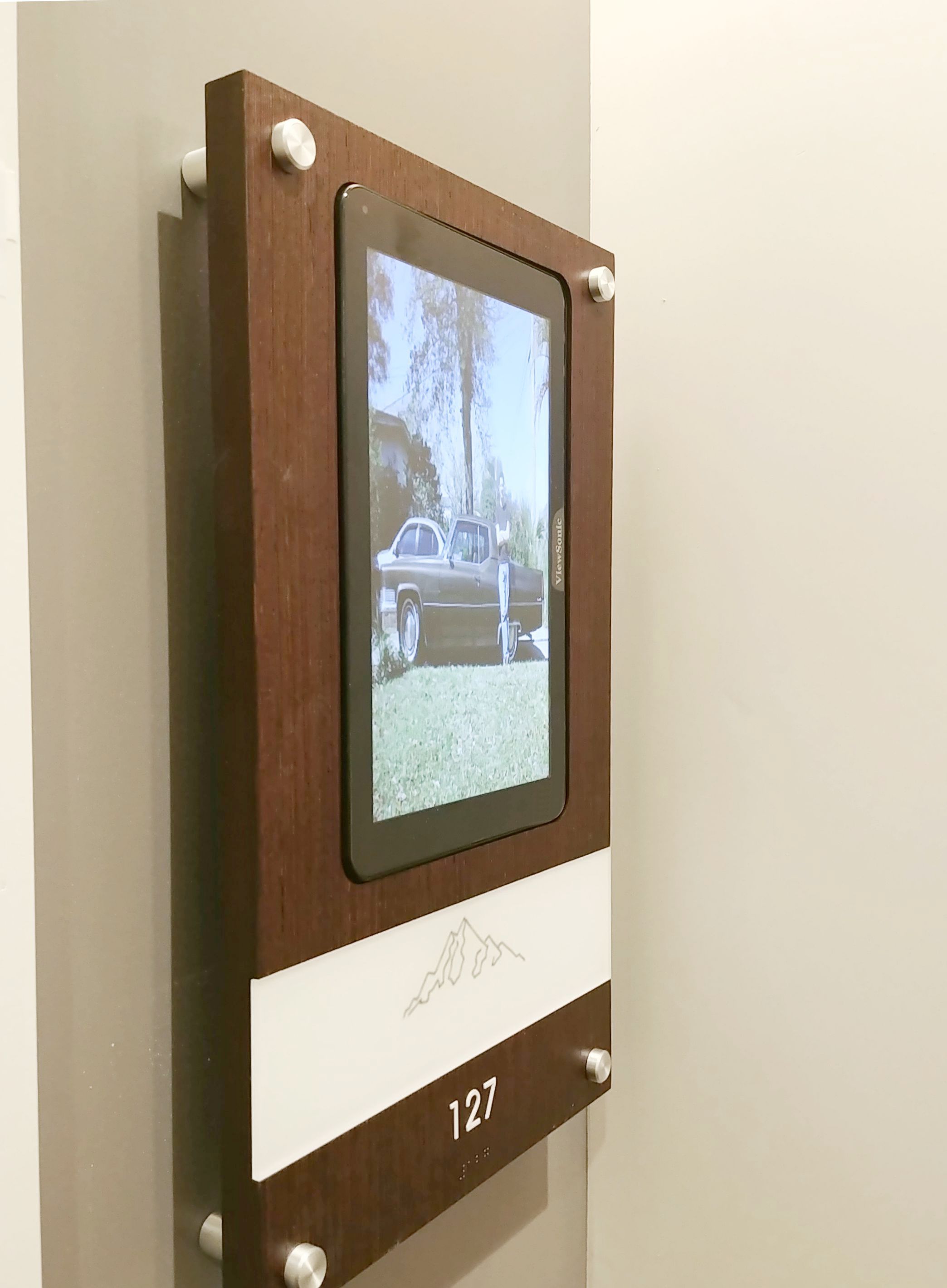 A digital photo frame displaying an image of a vintage car on grass is mounted on a beige wall. The frame is within a dark brown wooden plaque with silver accents. Below the screen is an engraving of a mountain range and the number "127" in white text.