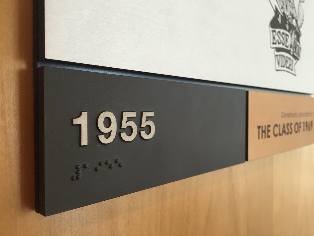 Close-up of a sign displaying the  number "1955" in raised metallic numbers with Braille text below. The sign is mounted on a wooden surface with adjacent text, partially visible, reading "THE CLASS OF 1969." The right upper corner has a partial graphic logo.