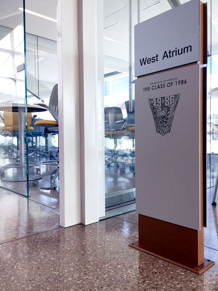 A modern hallway with floor-to-ceiling glass walls on the right, leading to a sunlit atrium labeled "West Atrium" on a sign. Inside the glass-walled room are high chairs and a table, with a polished concrete floor extending through the area.