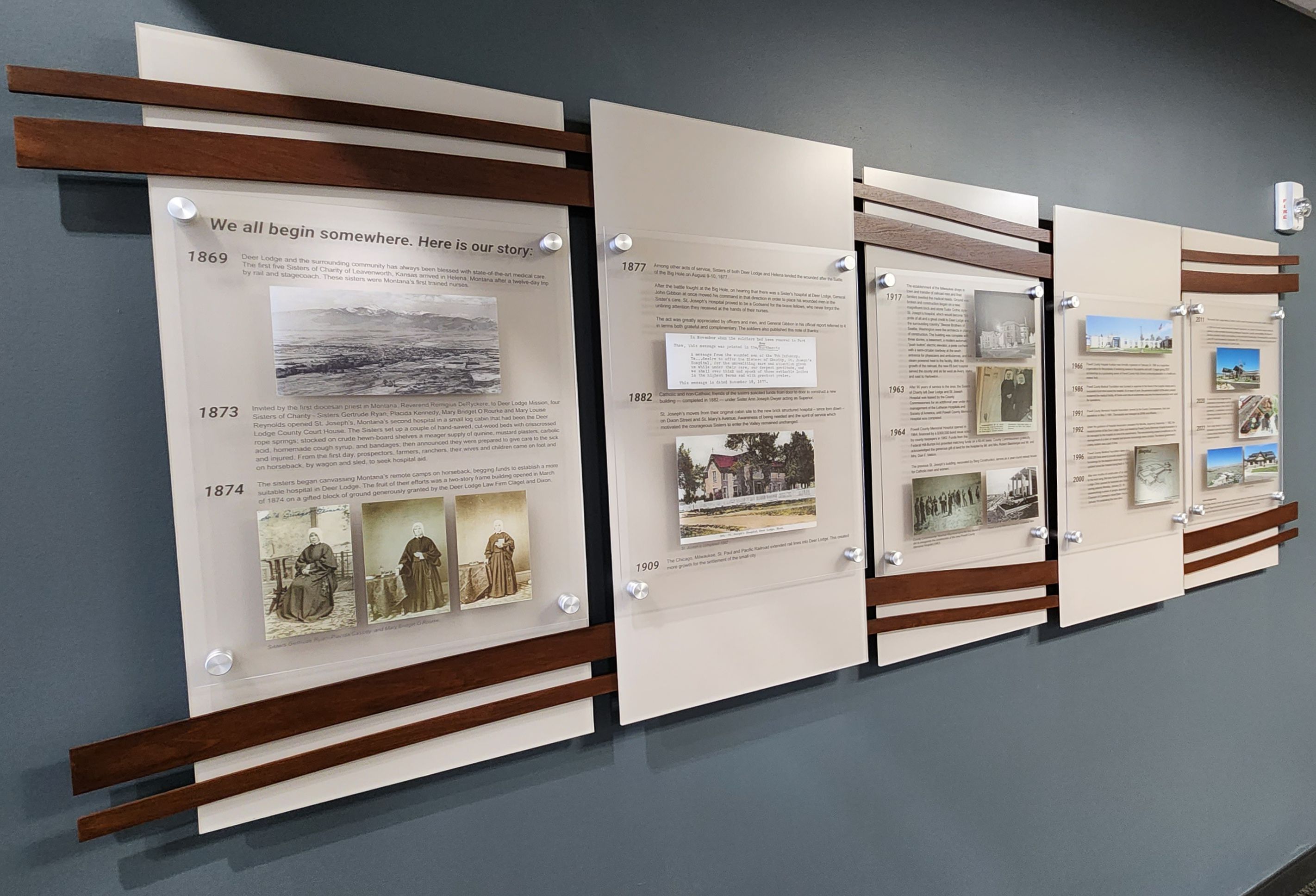 A display on a dark-colored wall features four acrylic panels with historical text, images, and dates ranging from 1869 to 1920. Each panel is bordered by wooden slats, and the first panel caption reads, "We all begin somewhere. Here is our story.