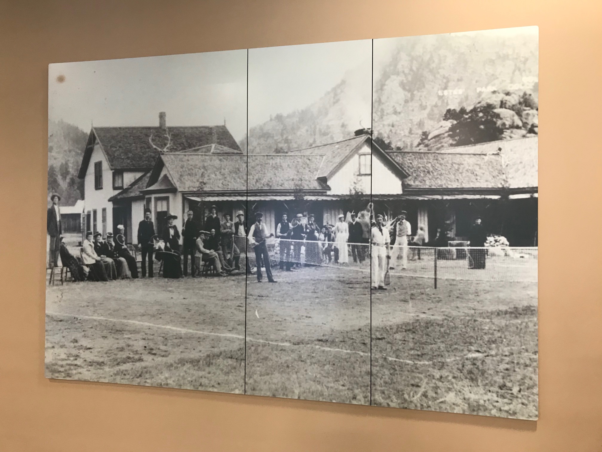 A large black-and-white historical photograph hangs on a light beige wall. The photo depicts a group of people standing outside old buildings.