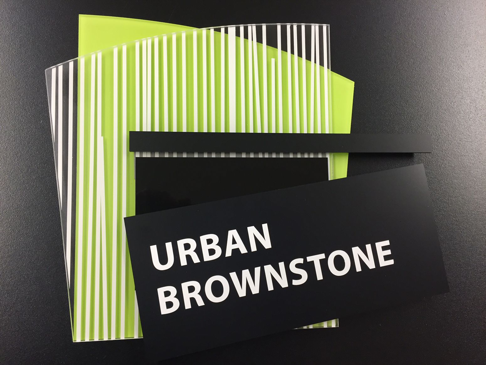 A black card with the text "URBAN BROWNSTONE" in white, placed on top of a stack of variously sized green and white striped translucent sheets. A black horizontal strip lies beneath the card, all set against a black background.
