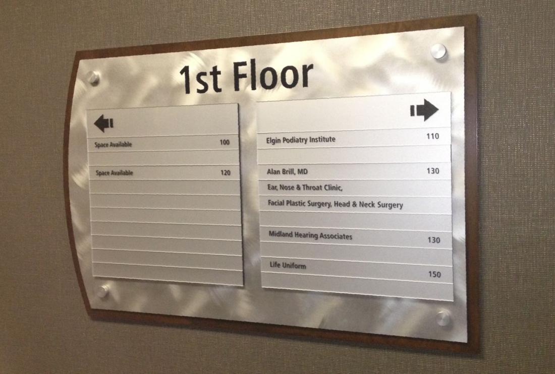 A metallic directory sign mounted on a wall indicates locations on the 1st floor. Listings include Elgin Podiatry Institute at 110, Alan Bill, MD at 130, ENT, Nose & Throat Clinic at 130, Facial Plastic Surgery at 130, Midland Hearing Associates at 140, and Life Uniform at 150.