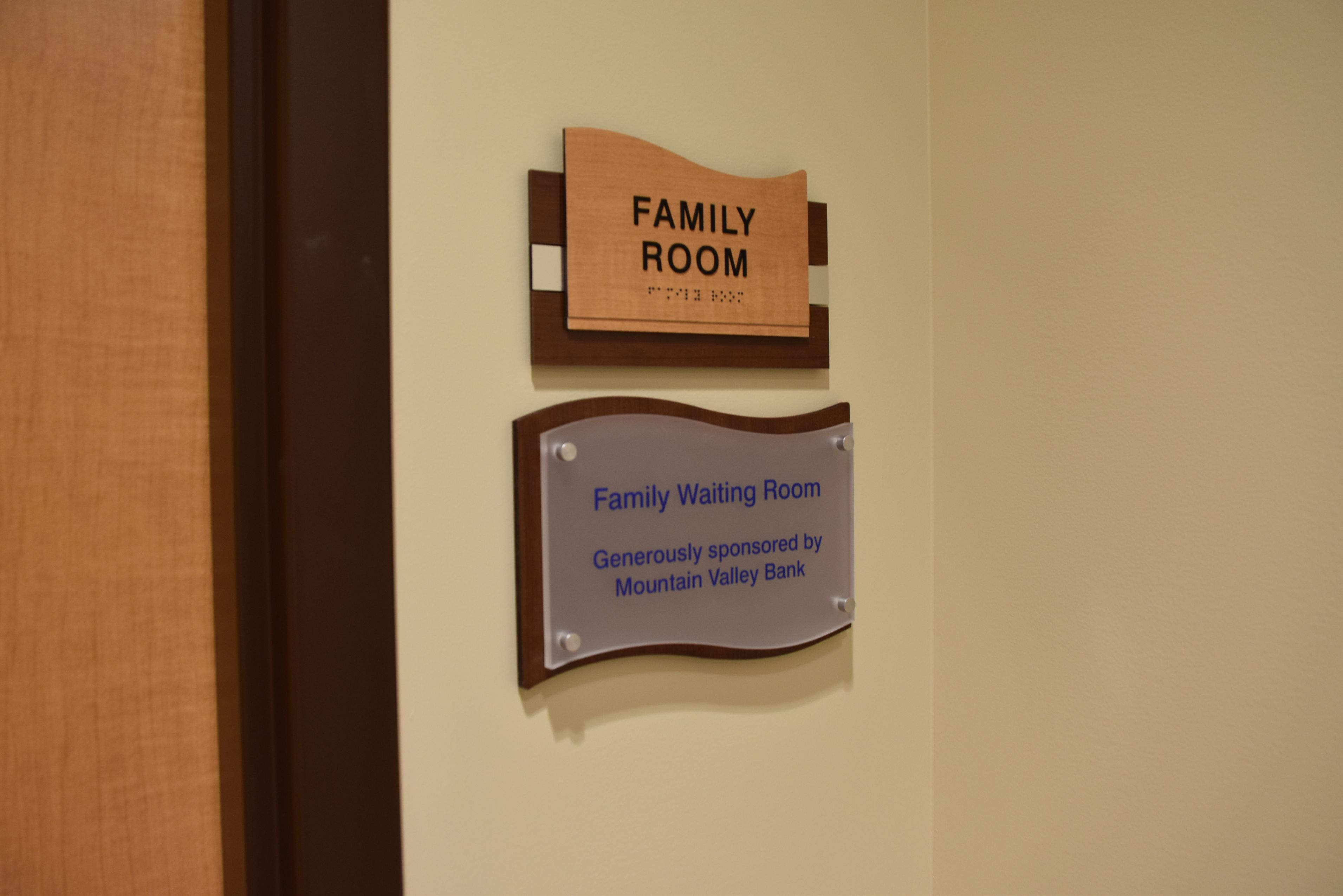 A wall sign reading "Family Room" is mounted above another sign that reads "Family Waiting Room Generously sponsored by Mountain Valley Bank." Both signs are displayed on a beige wall near a wooden door.