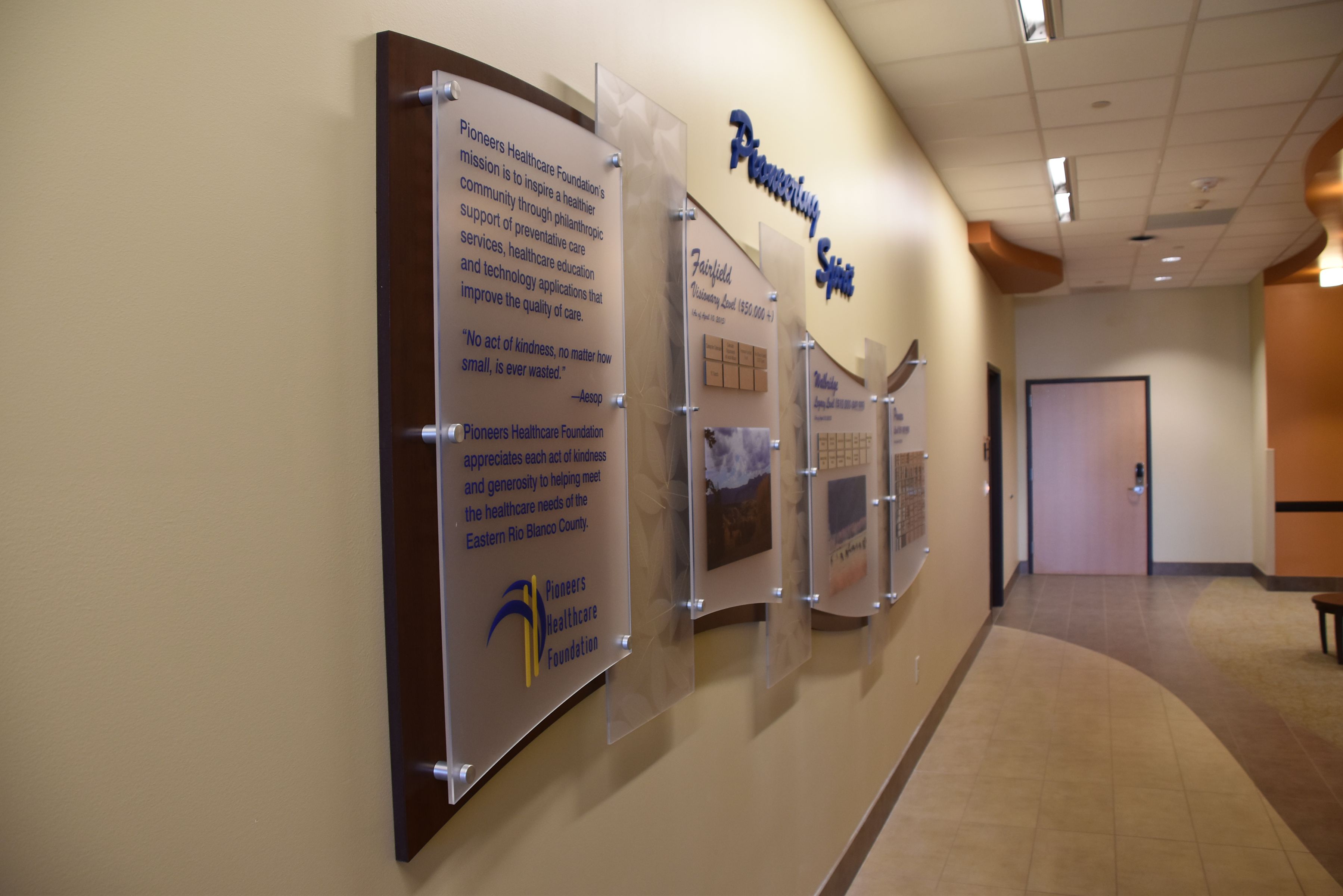A beige hallway with informational wall displays featuring text and images about a healthcare foundation. The displays are mounted on frosted acrylic panels with wooden backing. The ceiling has recessed lighting and a curved design. A door is visible down the hall.