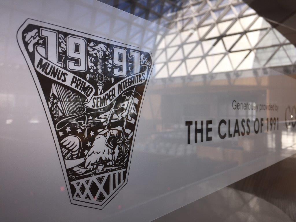 Close-up of a "Class of 1991" emblem on a glass panel. The emblem features mountains, trees, an eagle, and the Latin phrases "Munus Primo" and "Semper Integritas." The background shows a large, modern building with a glass roof structure.