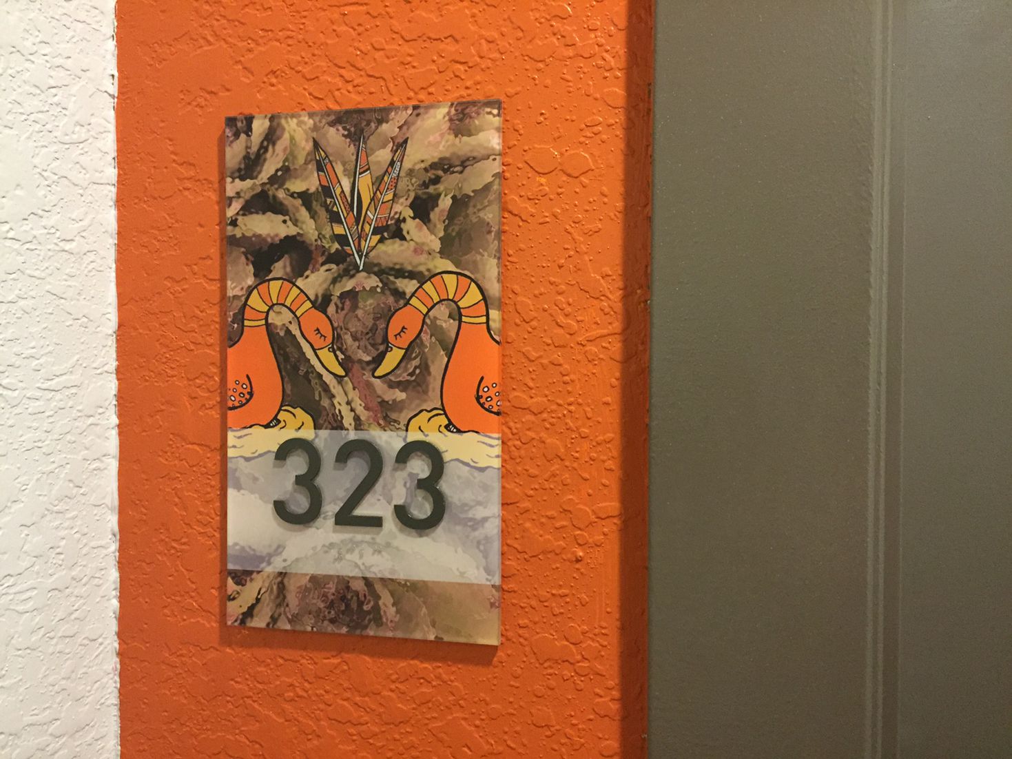 A decorative room number sign on an orange textured wall displays the number "323." The sign features a vibrant design with two mirrored orange birds and colorful feathers in the background.